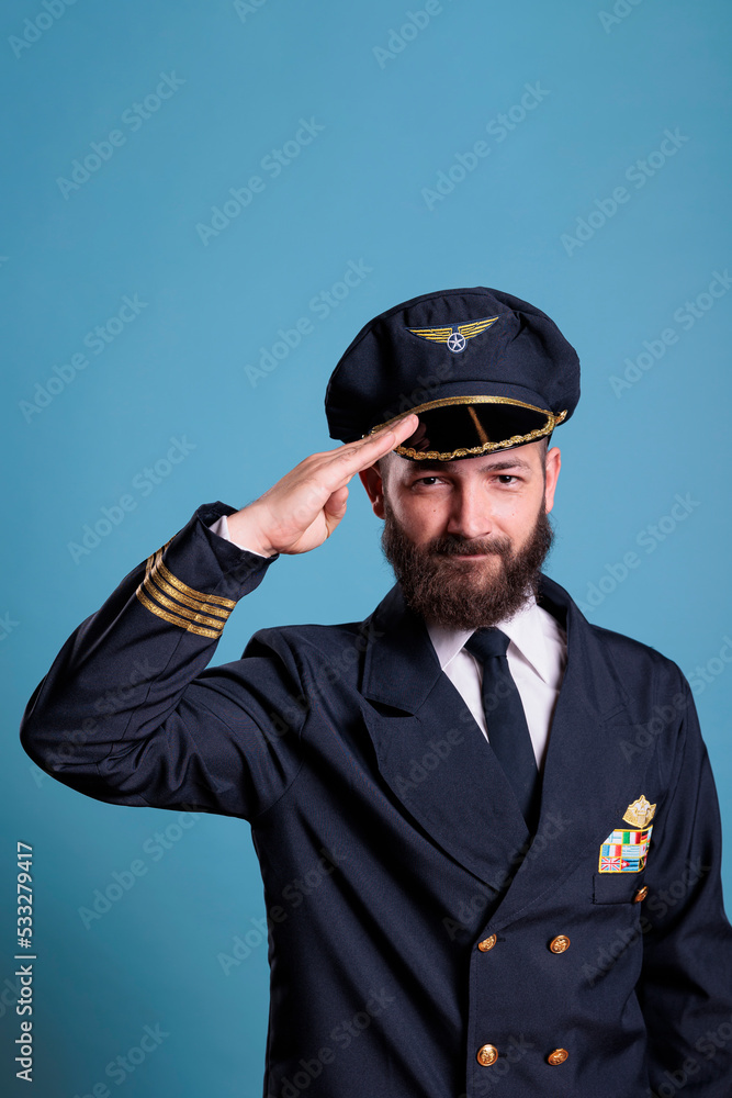 Smiling plane pilot saluting, wearing uniform and hat front view portrait, airplane captain looking at camera. Civil aviator with badge on jacket, aircrew member on blue background Smiling plane pilot saluting, wearing uniform and hat front view portrait, airplane captain looking at camera. Civil aviator with badge on jacket, aircrew member on blue background