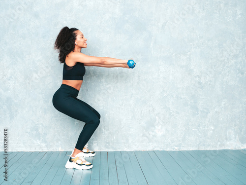 Fitness smiling black woman in sports clothing with afro curls hairstyle.She wearing sportswear. Young beautiful model doing squats. Female holding dumbbells in studio near grey wall