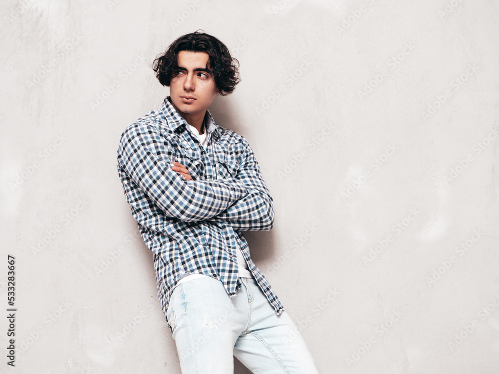 Portrait of handsome confident model. Sexy stylish man dressed in checkered shirt and jeans. Fashion hipster male posing near grey wall in studio. Crossed arms Portrait of handsome confident model. Sexy stylish man dressed in checkered shirt and jeans. Fashion hipster male posing near grey wall in studio. Crossed arms