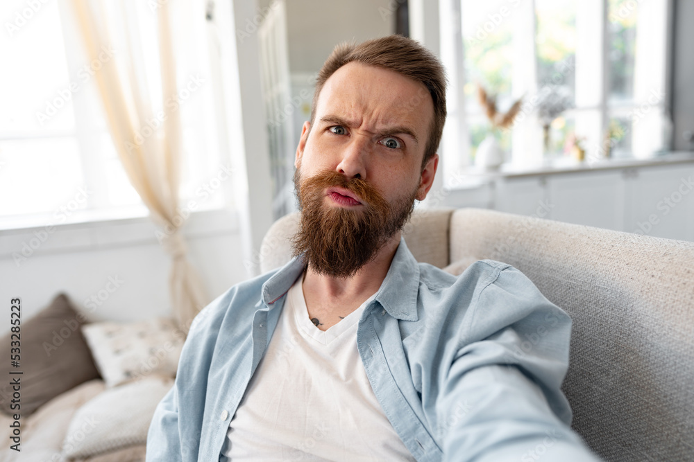 Bearded happy young man smiling and taking selfie photo while sitting on sofa at home. Bearded happy young man smiling and taking selfie photo while sitting on sofa at home.
