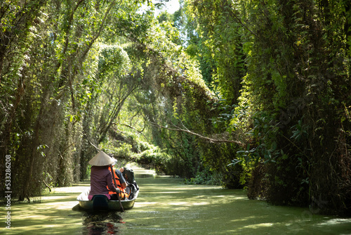 A group of tourists and local guide paddle a canoe along a beautiful canal with overhanging trees and vines in the Mekong Delta, Vietnam on a sunny day. Landscape format