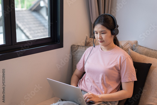 Front view, high angle shot portratit of middle-aged Asian woman in headset sitting on couch near windows, working, using laptop computer, smiling. Work at home, online study, technology concept. photo