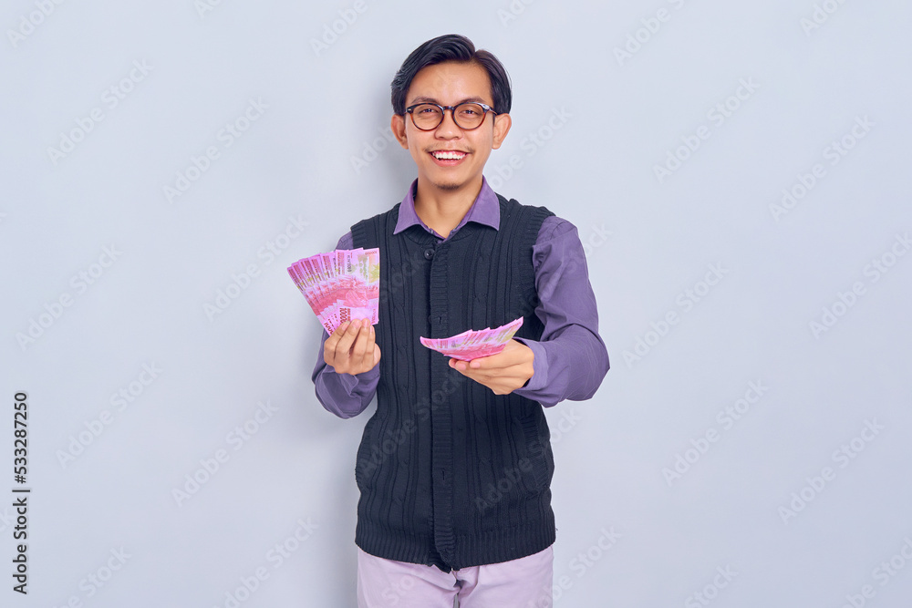 Portrait of smiling young handsome Asian man in shirt clothes giving cash money in rupiah banknotes to someone isolated on white background. People lifestyle concept Portrait of smiling young handsome Asian man in shirt clothes giving cash money in rupiah banknotes to someone isolated on white background. People lifestyle concept