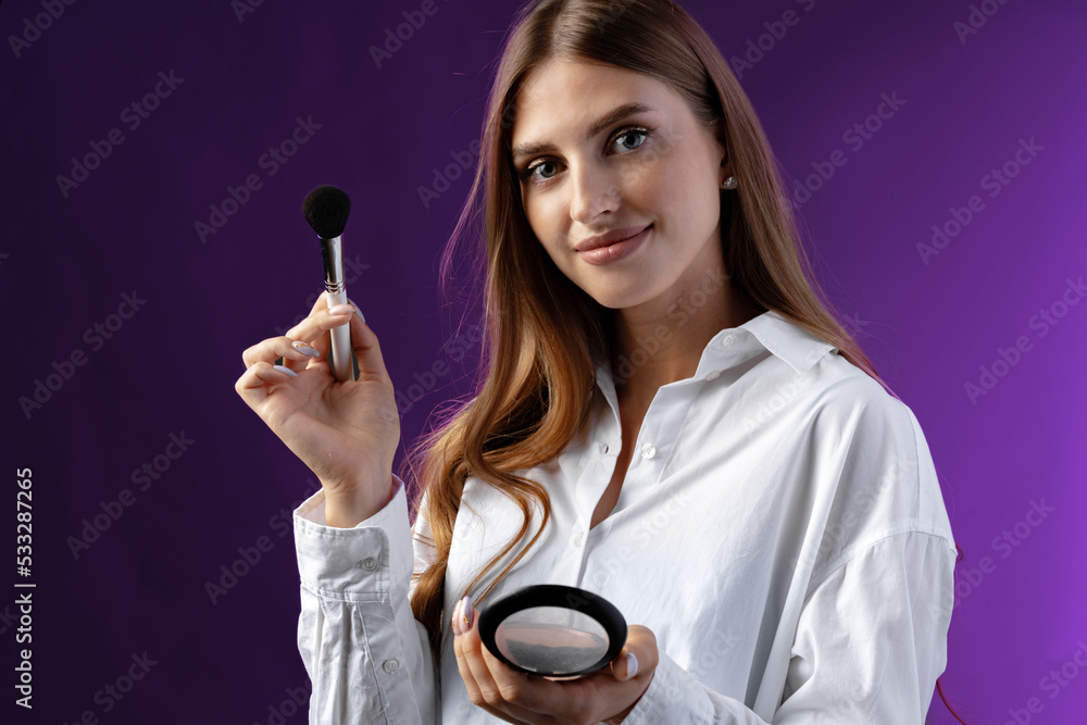 Young woman with makeup holding powder brush against purple background Young woman with makeup holding powder brush against purple background