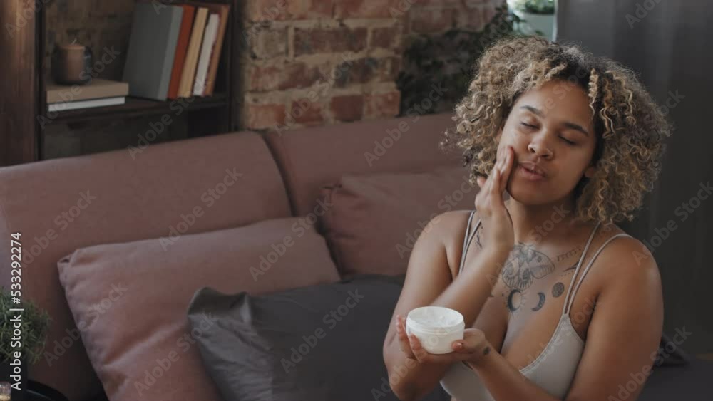 Modern young Black woman sitting on bed in loft bedroom applying moisturizing or nourishing cream over her face skin Modern young Black woman sitting on bed in loft bedroom applying moisturizing or nourishing cream over her face skin