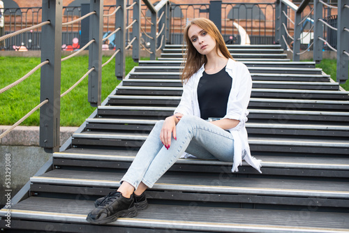 Portrait of a young and attractive Caucasian girl in casual clothes who is sitting on the steps in the park. © Павел Костенко