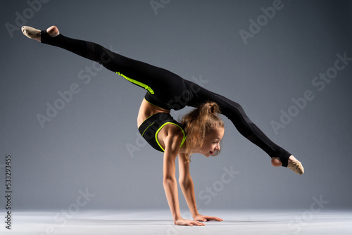 Flexible little girl doing acrobatic stunts while standing on her hands. The gymnast is dressed in a black tracksuit for stretching. Isolated on a gray background.