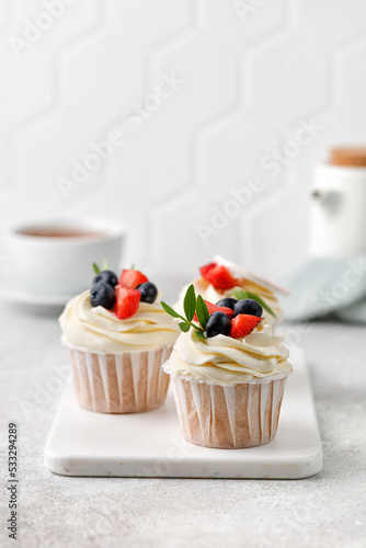 Vanilla cupcakes with fresh berries, buttercream, green leaves on grey background. Delicious homemade dessert. Copy space.
