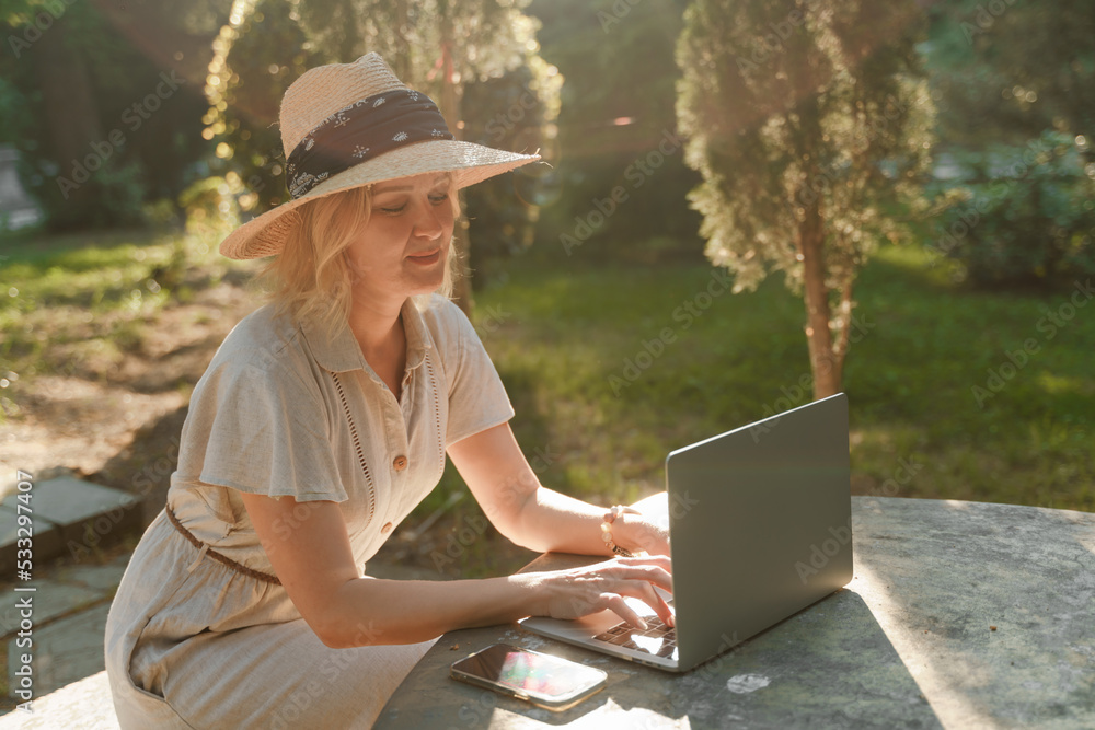 a beautiful mature woman in hat works on a computer at a white table in nature and spends her day productively