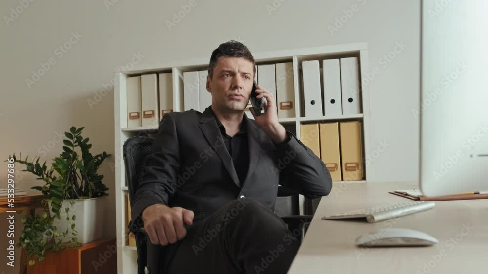 Medium shot of handsome young businessman in black formalwear making phone call while sitting at his workplace in modern office Medium shot of handsome young businessman in black formalwear making phone call while sitting at his workplace in modern office
