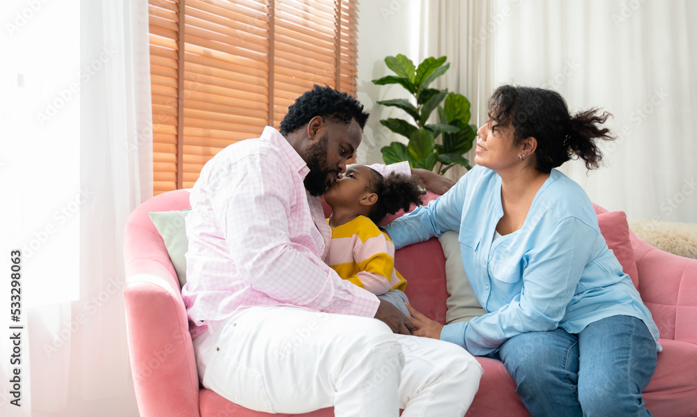Happy family relaxing together on sofa at home. African little girl kissing her dad on cheek. . Happy family relaxing together on sofa at home. African little girl kissing her dad on cheek. .