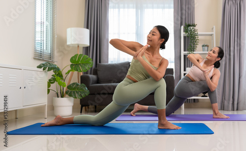 Two women doing yoga exercise at home, stretching in Parivrtta Anjaneyasana or Revolved Low Lunge Pose in  bedroom. Working out wearing sportive top and leggings. Relaxation and Healthcare concept.. photo