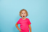 Portrait of shocked little girl with curly fair hair wearing pink T-shirt, looking with open mouth on blue background.