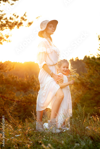 Side view of gorgeous family wearing white dresses, standing near pines in forest lit up by sunset in summer. Vertical.