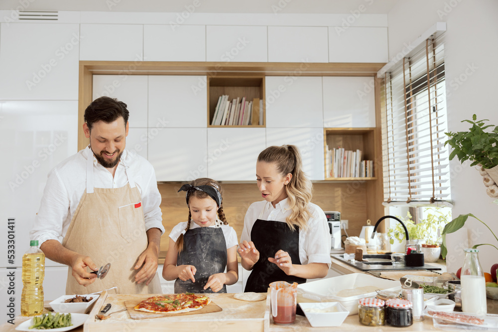 Delighted surprised kid and mother waiting for eating homemade domestic pizza. Father jolding pizza cutter cutting pizza. Happy family preparing cooking baking dinner in modern kitchen with window. Delighted surprised kid and mother waiting for eating homemade domestic pizza. Father jolding pizza cutter cutting pizza. Happy family preparing cooking baking dinner in modern kitchen with window.