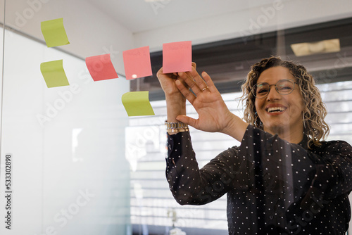 Happy mature businesswoman writing on sticky notes on glass at office photo
