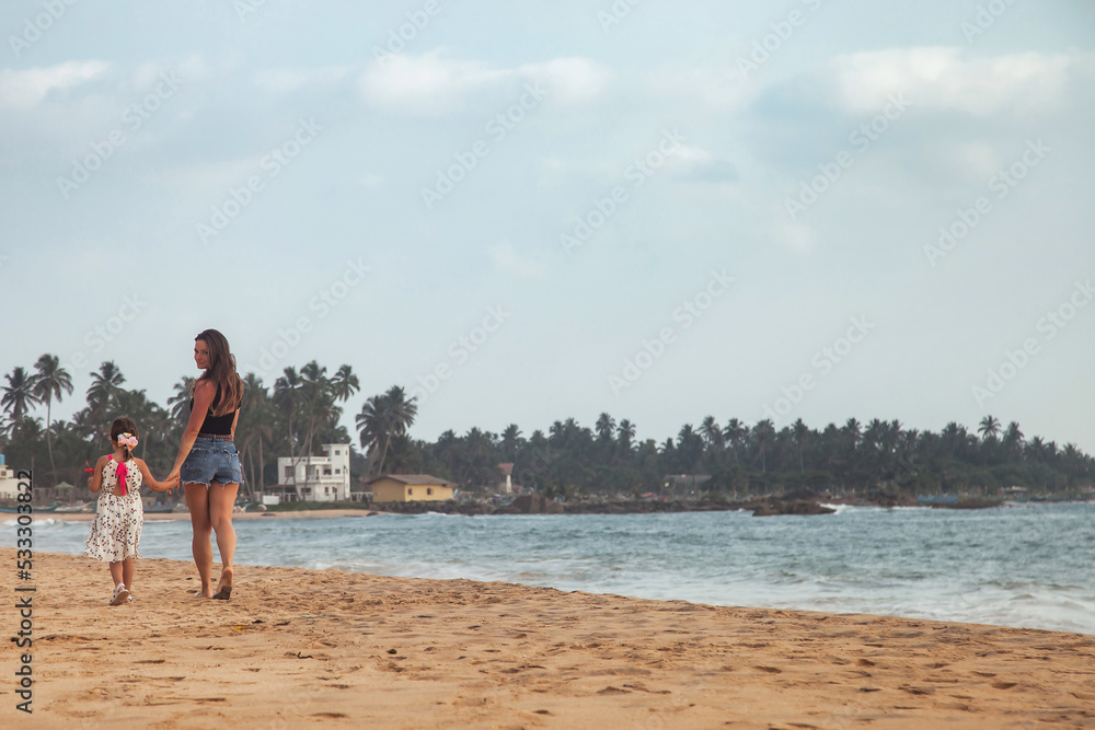 Rear view mom and little daughter together walking on tropical sandy beach at sea background. Mother and little girl in casual clothes on ocean. Concept family vacation, travel. Copy text space