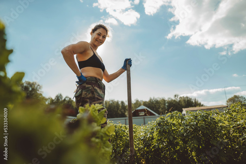 Smiling mature woman holding shovel standing with hand on hip in vegetable garden photo