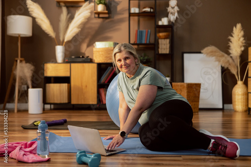 Senior woman granny sitting on yoga mat surfing in internet yoga exercise online. Blue dambbells and bottle with water on floor. Woman trains does exercises looks at camera smiling spending time.