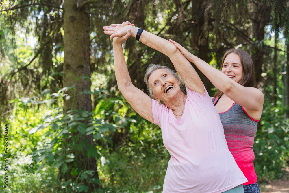 Happy fitness instructor guiding senior woman exercising in front of tree at park Happy fitness instructor guiding senior woman exercising in front of tree at park