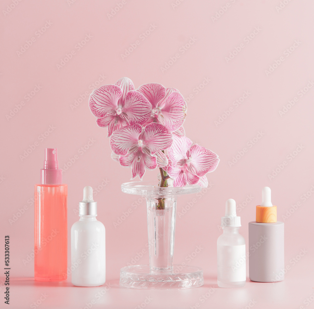 Modern cosmetic products setting with various bottles and orchid flowers at pastel pink background. Front view. Beauty concept Modern cosmetic products setting with various bottles and orchid flowers at pastel pink background. Front view. Beauty concept
