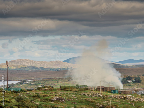 Tall white smoke rising into blue cloudy sky from a farm in rural area. Stunning nature scenery in the background. West of Ireland. Farm accident concept. Warm sunny day.