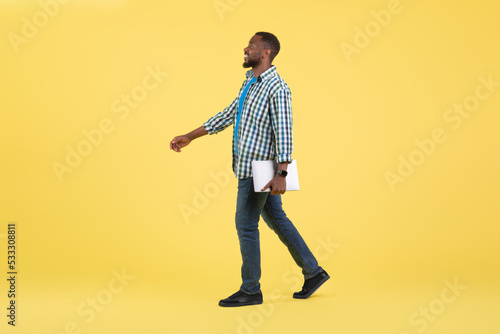 Happy Black Guy Holding Laptop Walking Over Yellow Background