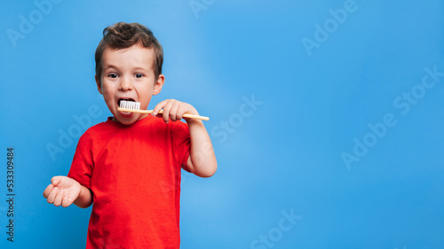 Smiling boy with healthy teeth brushing his teeth with a toothbrush on a blue isolated background. Oral hygiene. A place for your text.