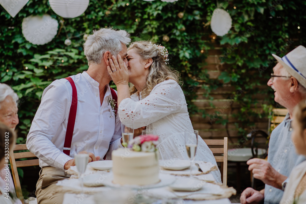 custom made wallpaper toronto digitalMature bride and groom kissing at wedding reception with their family, outside in the backyard.