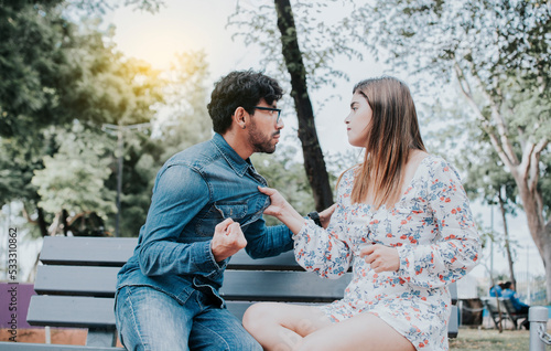 Upset couple arguing on a park bench. Young couple arguing sitting on a park bench  Concept of aggressive couples in the park. Man arguing with his girlfriend sitting in a park