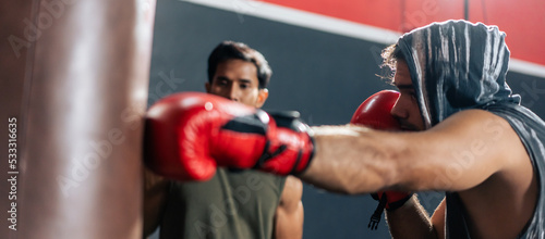 Asian man wearing boxing gloves punching ahead with trainer in fitness © Kawee