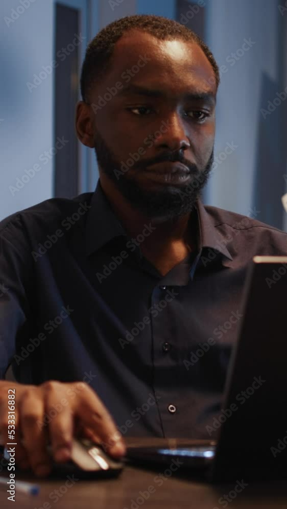 Vertical video: African american man working on laptop in startup office, analyzing data on internet and documents late at night. Using research to create report and presentation with app. Handheld Vertical video: African american man working on laptop in startup office, analyzing data on internet and documents late at night. Using research to create report and presentation with app. Handheld