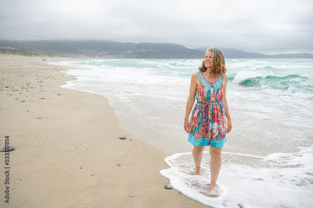 An adult woman walks along an empty and wild beach with waves from the sea.