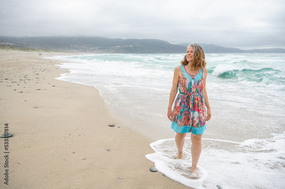 An adult woman walks along an empty and wild beach with waves from the sea. An adult woman walks along an empty and wild beach with waves from the sea.