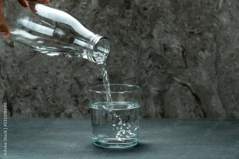 Detail of the hand of an unrecognizable man pouring water from a glass bottle into a glass on a textured background. Detail of the hand of an unrecognizable man pouring water from a glass bottle into a glass on a textured background.