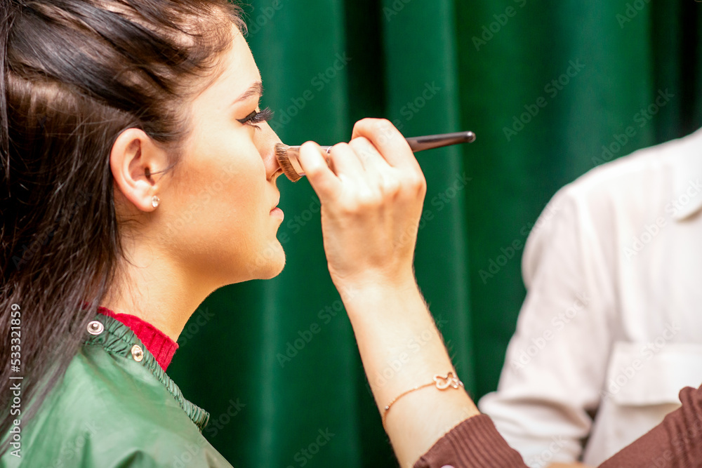 Obraz premium The face of the young woman receives powder on her nose with a brush with the hand of a makeup artist