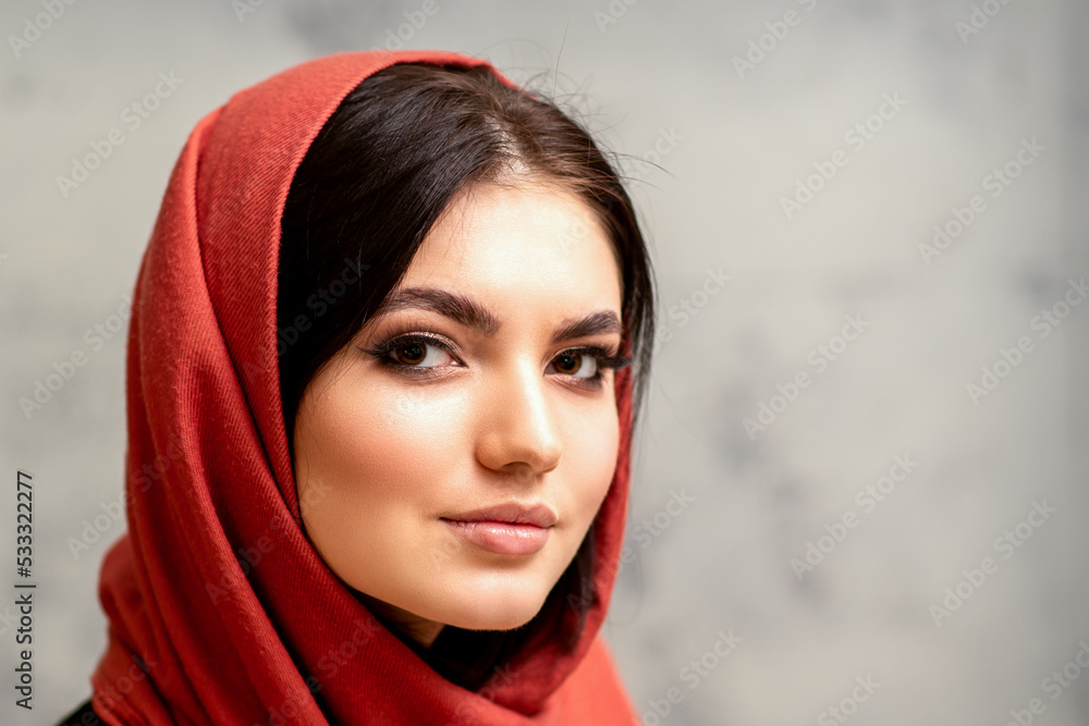 The fashionable young woman. Portrait of the beautiful female model with long hair and makeup in a red scarf. Beauty young caucasian woman on the background of a gray wall The fashionable young woman. Portrait of the beautiful female model with long hair and makeup in a red scarf. Beauty young caucasian woman on the background of a gray wall