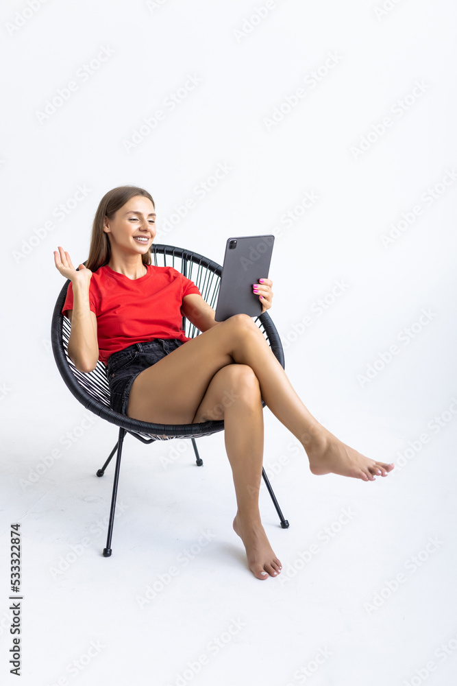 Portrait of cheerful young woman with digital tablet sitting on chair, working online against white studio wall, free space. Modern technologies and communications concept Portrait of cheerful young woman with digital tablet sitting on chair, working online against white studio wall, free space. Modern technologies and communications concept