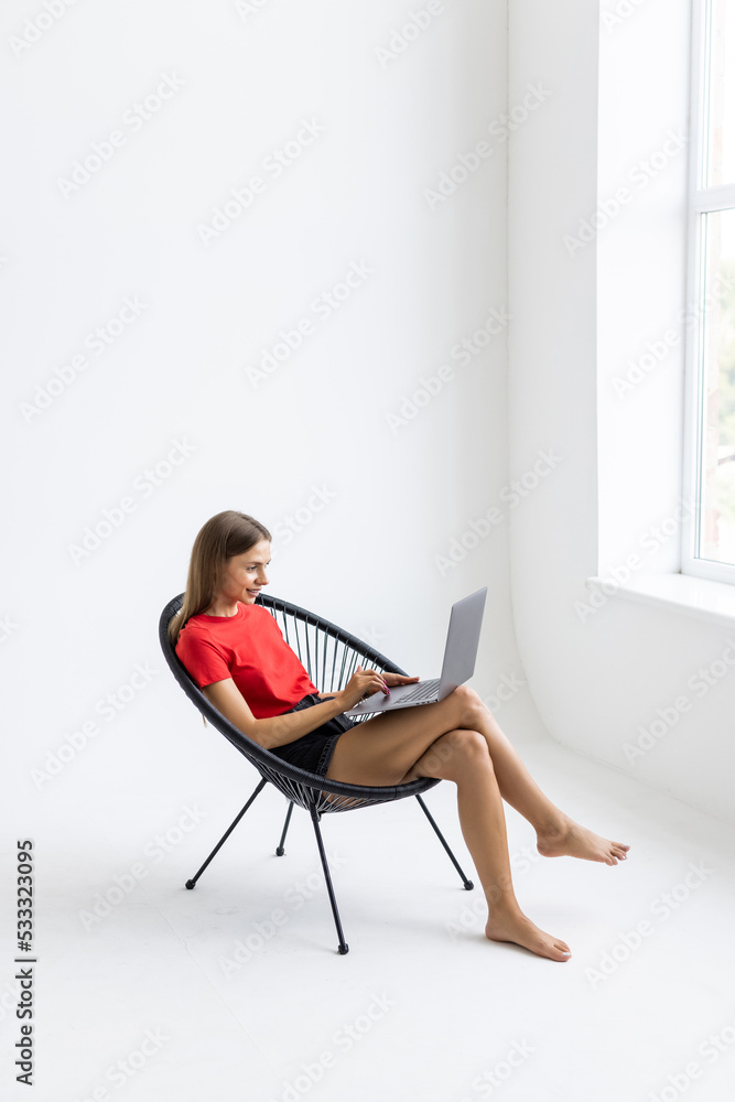 Young smiling woman sitting on modern chair near the window in light cozy room at home working on laptop in relaxing atmosphere Young smiling woman sitting on modern chair near the window in light cozy room at home working on laptop in relaxing atmosphere