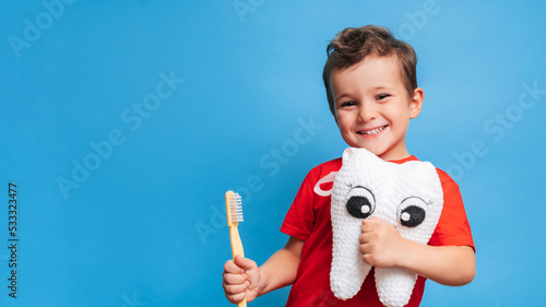 A smiling boy with healthy teeth holds a plush tooth and a toothbrush on a blue isolated background. Oral hygiene. Pediatric dentistry. Prevention of caries. A place for your text. photo