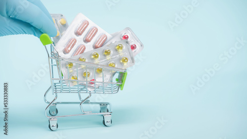 cropped view of person in latex glove holding small shopping cart with pills on blue background.