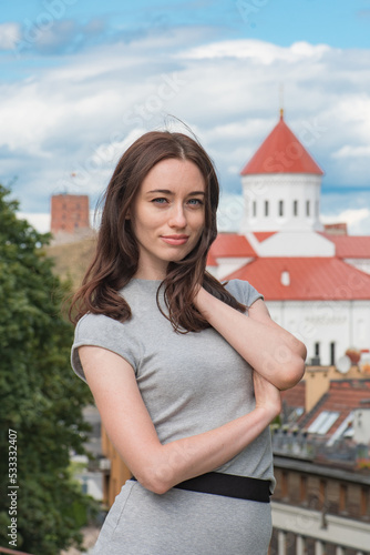 Beautiful Ukrainian caucasian girl with red hair, blue eyes and freckles in a grey dress posing and smiling with Gediminas castle and a church in Vilnius old town on background, vertical