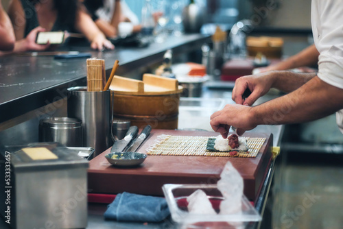 Close-up of a chef's hands rolling a sushi roll with rice and seaweed at a sushi bar with people in the background at the counter
