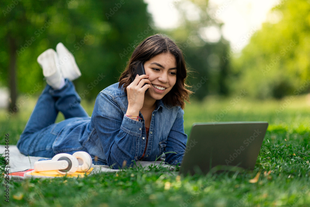 Smiling Middle Eastern Female Student Relaxing On Lawn With Laptop And Cellphone Smiling Middle Eastern Female Student Relaxing On Lawn With Laptop And Cellphone