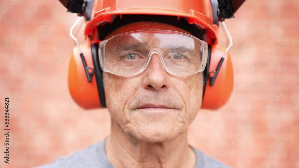 Portrait closeup of a mature man worker with a safety helmet, headphones, and protective goggles looking at the camera and smiling. Portrait closeup of a mature man worker with a safety helmet, headphones, and protective goggles looking at the camera and smiling.