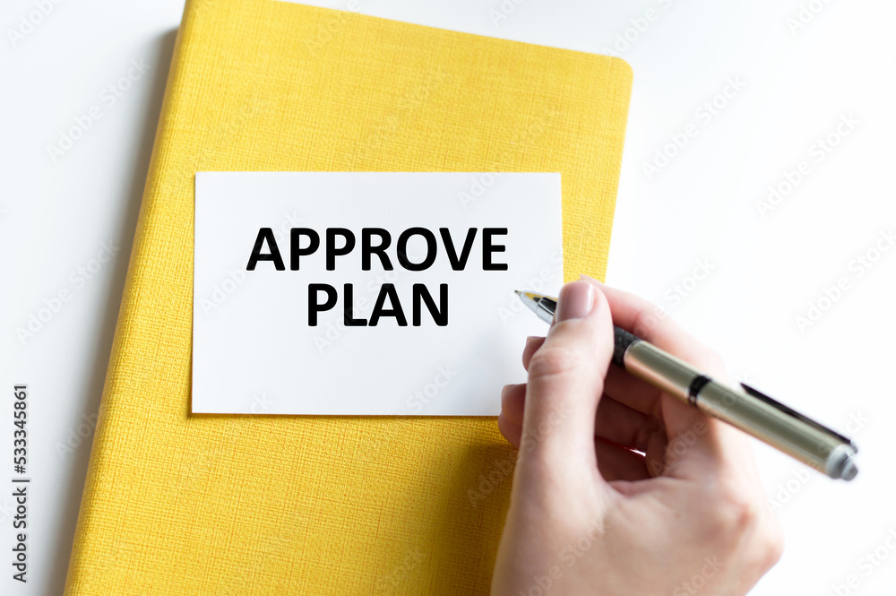 Text APPROVE PLAN on a card with a yellow notepad next to a hand with a person's pen Text APPROVE PLAN on a card with a yellow notepad next to a hand with a person's pen