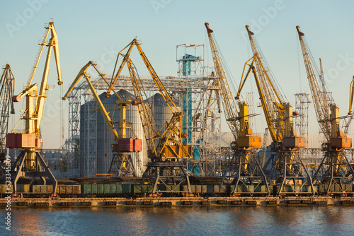 Many yellow port cranes are working in the port, in the background is a silver grain terminal