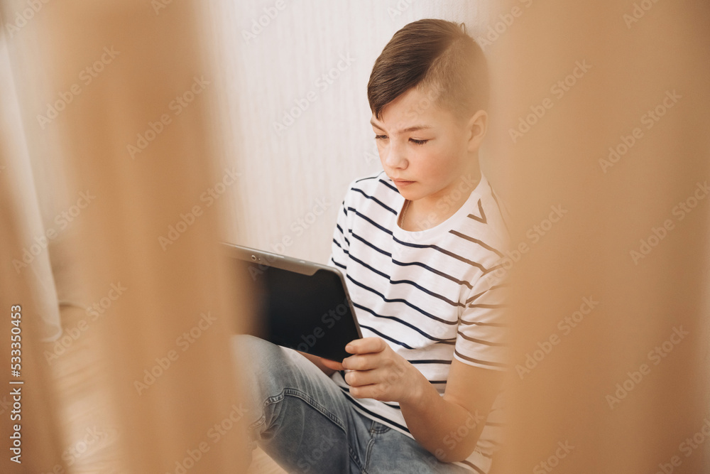 Teenage boy sitting on step of stairs using tablet playing video games on internet online at cozy home. Social media