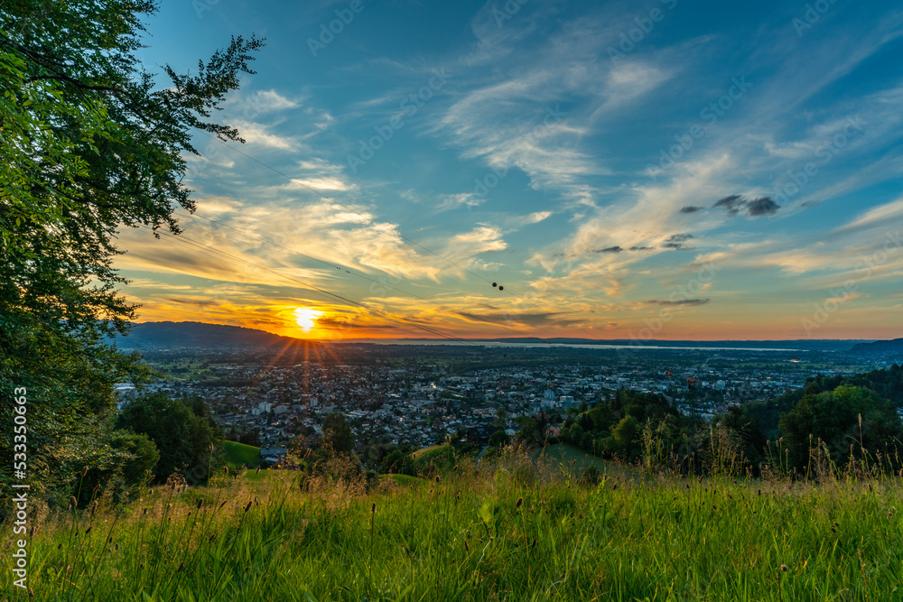 Sonnenuntergang über den Schweizer Bergen im Rheintal, Vorarlberg, Austria. leuchtendes Abendrot, tolle Stimmung mit roten und graublauen Wolken über den grünen Wiesen und Bäumen. Seilbahn zum Karren