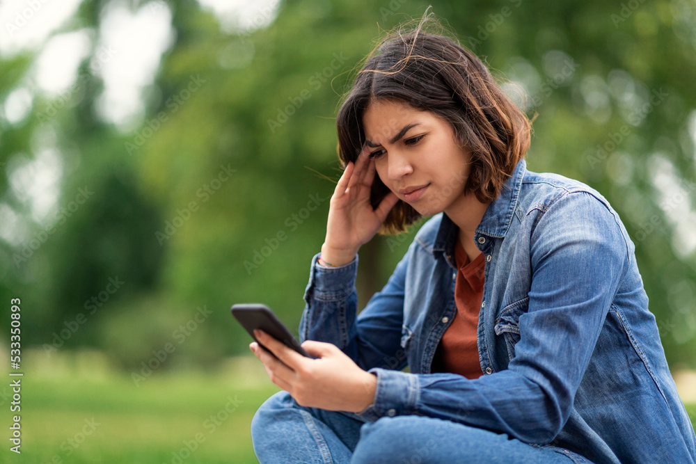 Worried Young Arab Female Reading Message On Smartphone While Sitting Outdoors